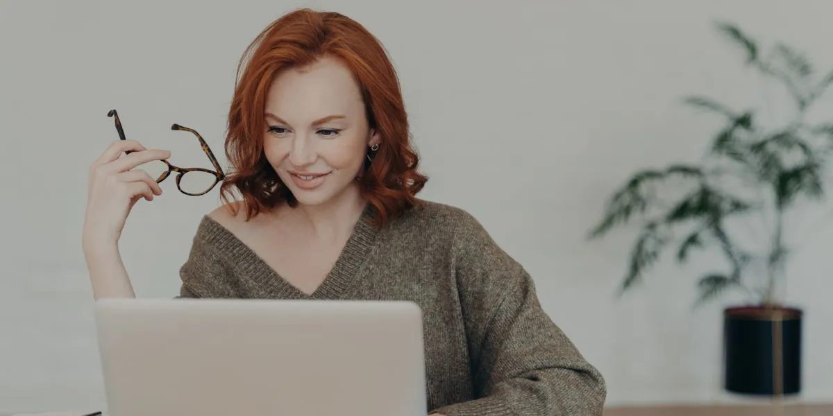 Femme rousse tenant ses lunettes en main vérifie un travail sur PC